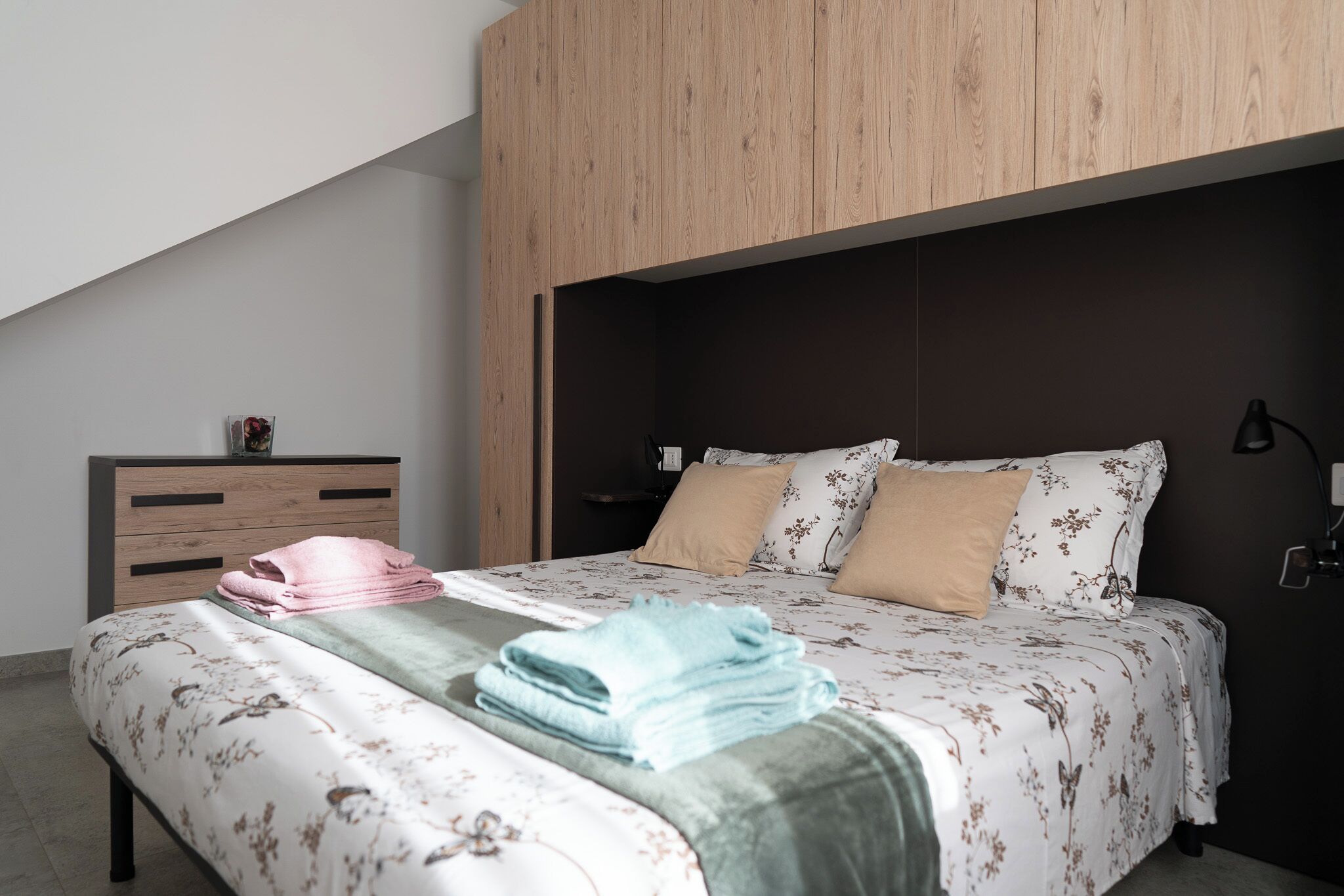 Une chambre à coucher avec une tête de lit en bois, un lit recouvert d’une literie à motifs floraux, une table de chevet avec une lampe et une petite table sur laquelle est posé un vase.