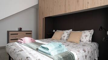 Une chambre à coucher avec une tête de lit en bois, un lit recouvert d’une literie à motifs floraux, une table de chevet avec une lampe et une petite table sur laquelle est posé un vase.