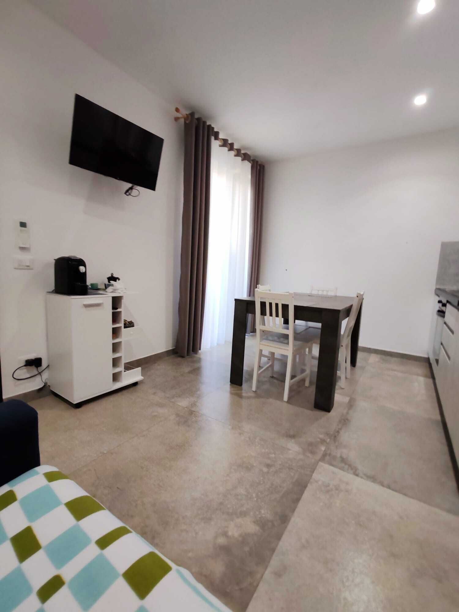 A small, clean room with a bed, a desk, a chair, a TV mounted on the wall, and a window with curtains.