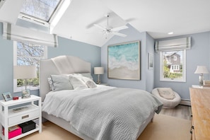 Une chambre à coucher comprenant un lit, des tables de chevet, un ventilateur de plafond, une fenêtre avec vue et un tableau accroché au mur.