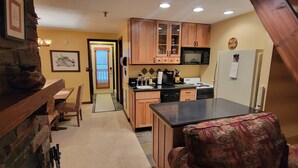 A compact kitchen with wooden cabinets, a microwave, a stove, and a refrigerator. There is a dining area with a table and chairs, and a patterned armchair. - Cozy Loft nestled atop of Snowshoe (Snowshoe)