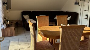 A dining area with a round table and wicker chairs, a sofa in the background, and a ceiling light fixture. - Vacation apartment Haren /Ems for up to 6 people  (Haren (Ems))