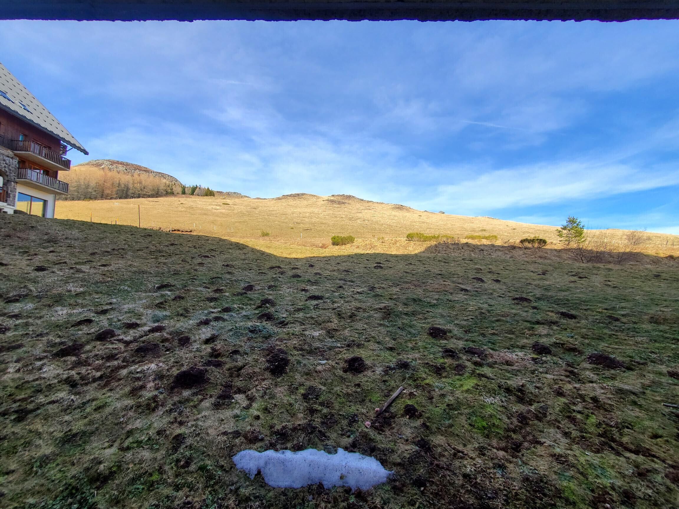 Eine Wiese mit einer kleinen Schneefläche, ein Holzbalkon und ein klarer Himmel.