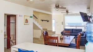 A spacious living area with a dining table, chairs, and a sofa. There are two ceiling fans, a staircase, and a large window offering a view of the sea.