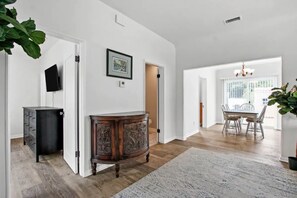 Una habitación amplia con piso de madera, un mueble de madera grande, una zona de comedor con mesa y sillas, y una araña.