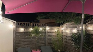 Um pátio externo com duas cadeiras de descanso, um guarda-sol rosa, luzes de Natal e um cercado de madeira.