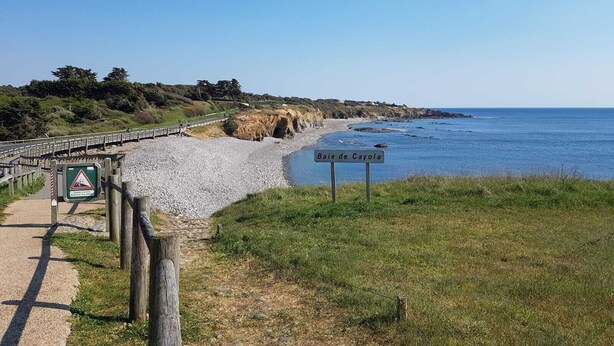 Um caminho costeiro com uma placa indicando "Baie de Cayola", uma cerca de madeira e uma praia rochosa.