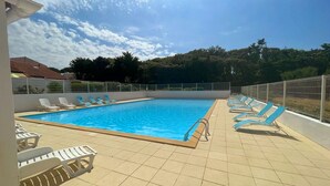 Piscina com espreguiçadeiras em deck de azulejos, cercada por uma grade de metal, sob céu claro.