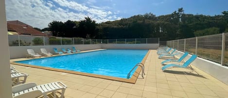 A swimming pool with sun loungers on a tiled deck, surrounded by a metal fence, under a clear sky.