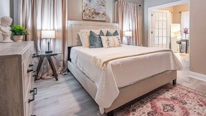 Een slaapkamer met een bed, gordijnen, een schilderij, een lamp, een dressoir en een plant.