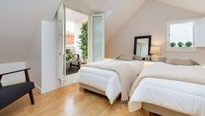 A bedroom with two beds, a desk, a chair, a window with a view, and a door leading to an outdoor area.