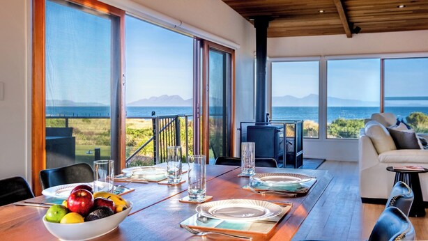 Un coin repas avec une table dressée pour quatre personnes, de grandes fenêtres offrant une vue sur la mer et les montagnes, et un espace salon confortable avec un canapé et une petite table.