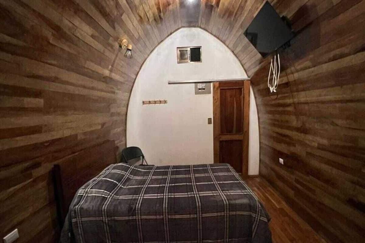 Une pièce aux murs en bois et au plafond en pente, avec un lit recouvert d’une couverture à carreaux, une petite fenêtre et une porte.