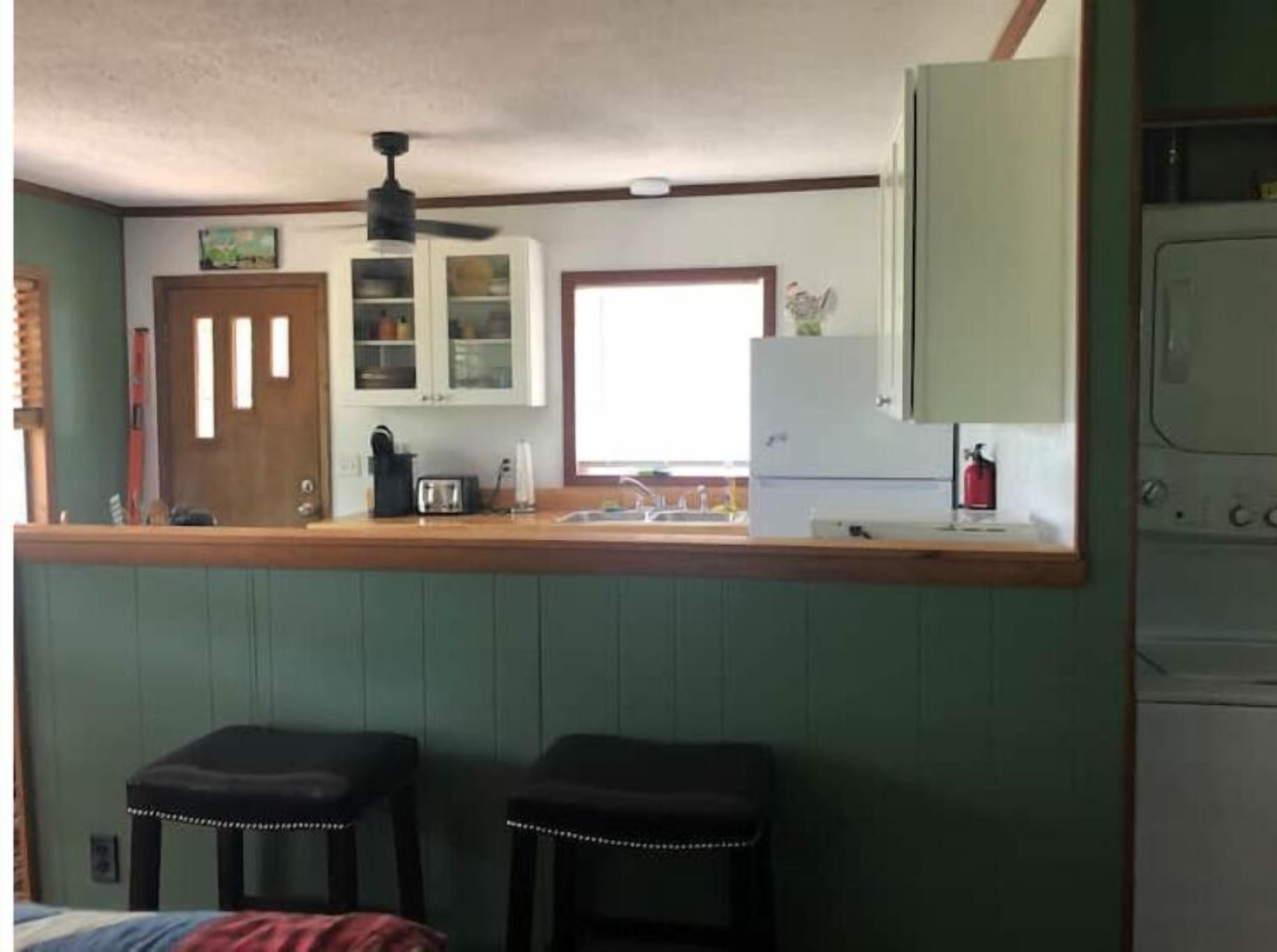 Una cucina compatta con un piano di lavoro in legno, una ventola a soffitto e un frigorifero. Ci sono due sgabelli al bancone e una finestra che lascia entrare luce naturale.