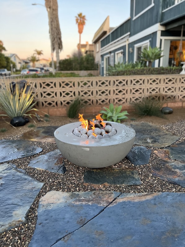 A fire pit with burning logs in a circular concrete design, surrounded by a stone pathway and various plants. - Seahorse Cottage Luxe (Oxnard)