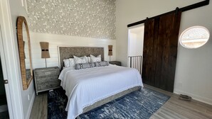 A bedroom with a large bed, a wooden headboard, a dark blue rug, and a wooden sliding door. - Seahorse Cottage Luxe (Oxnard)