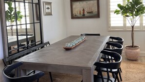 A dining area with a long table, black chairs, a woven light fixture, a large mirror, a framed picture, and a potted plant. - Seahorse Cottage Luxe (Oxnard)