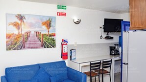 A room with a blue sofa, a desk with chairs, a TV, a fire extinguisher, and a painting of a beach scene.