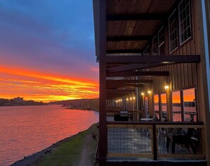 A waterfront building with a covered outdoor seating area and large windows overlooking a sunset over a body of water.
