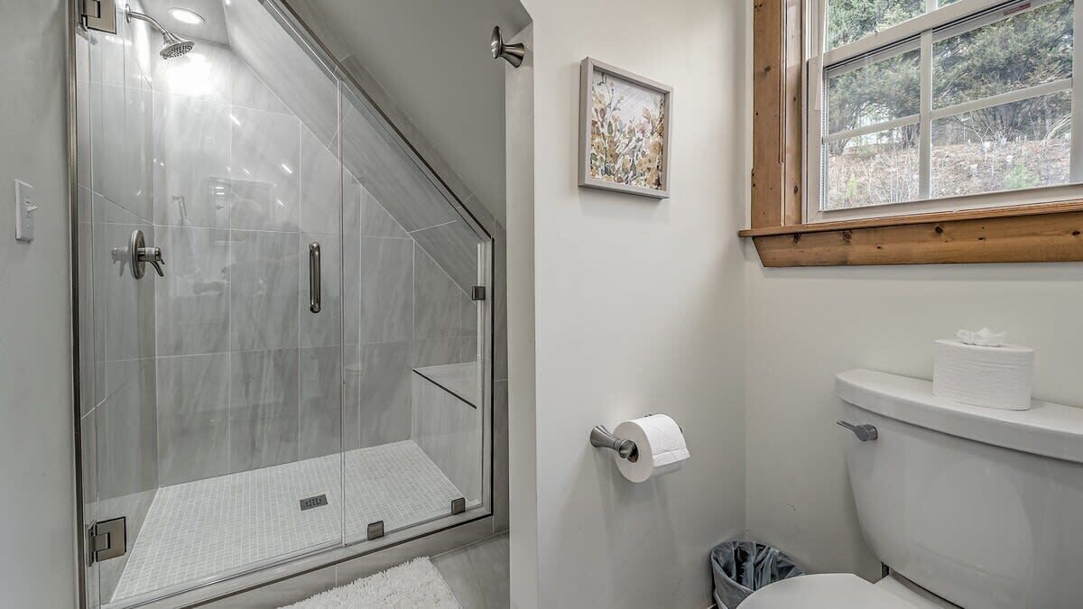 Bagno con doccia vetrata, un water, una finestra e un quadro appeso al muro.