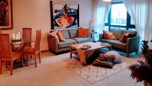 A modern living room with two sofas, a coffee table, and a dining area with chairs. There is a large painting on the wall, a lamp, and a window with curtains. - 9 mins from the northern line station. 2 mins from 3 B/stops and local amenities (London)