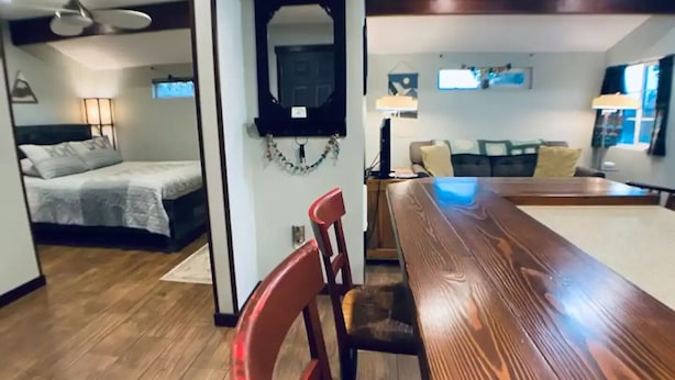 Un interior moderno con una mesa de comedor de madera, una silla roja, una cama con edredón blanco y un ventilador de techo.