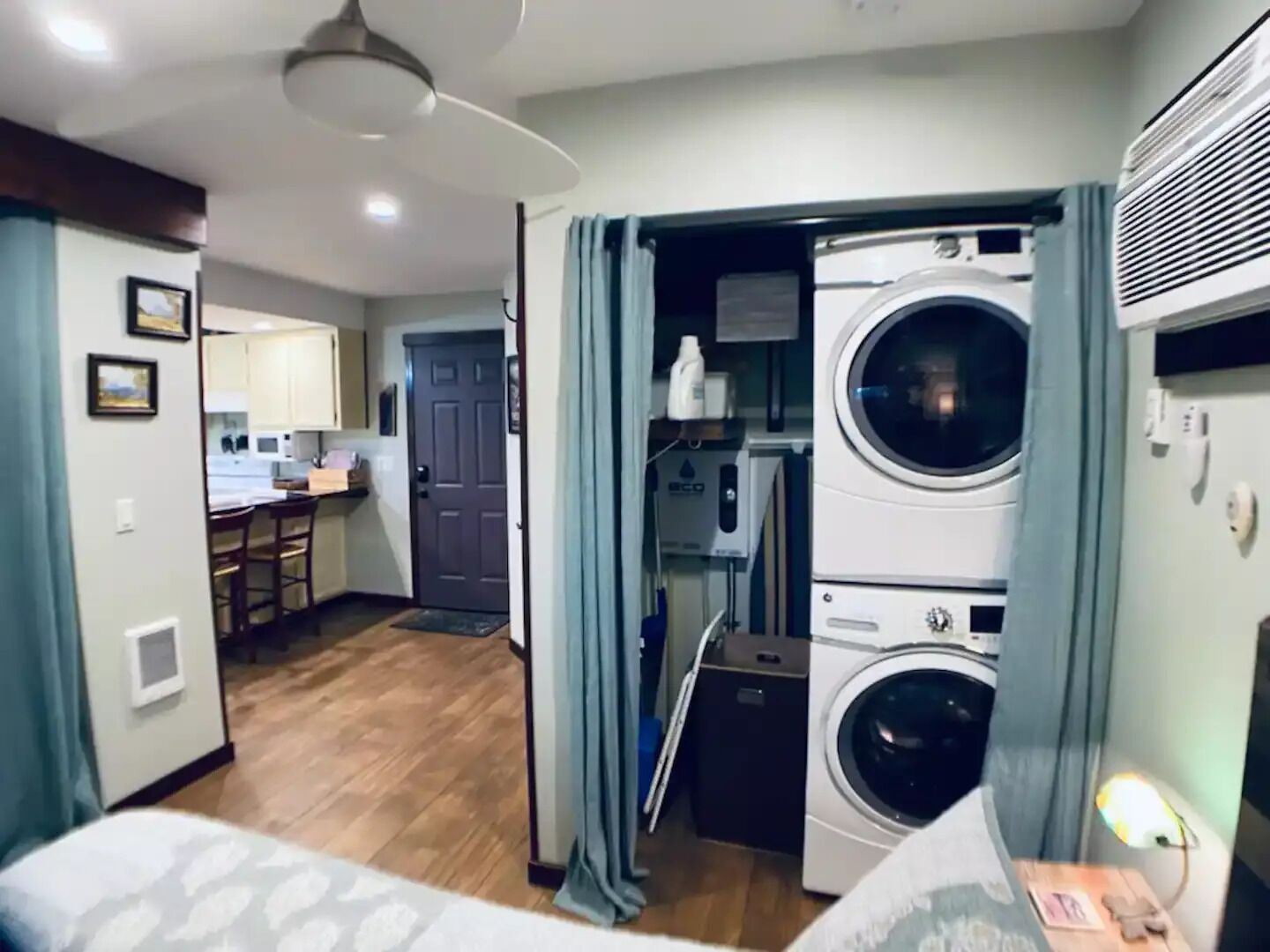 A compact hotel room with a bed, a small bedside table, a nightstand, and a view of a kitchen and laundry area.