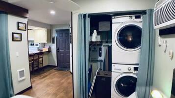 A compact hotel room with a bed, a small bedside table, a nightstand, and a view of a kitchen and laundry area.