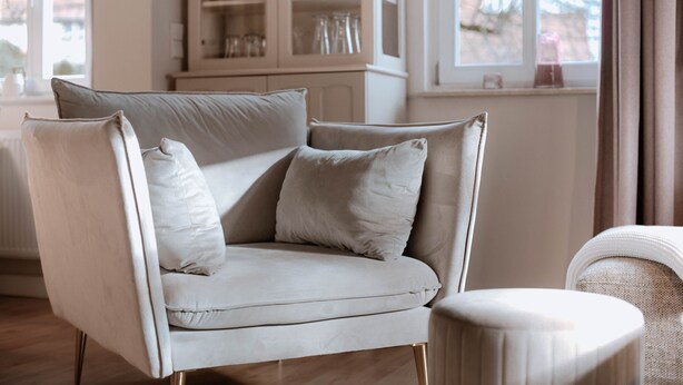 A modern living room with a light-colored sofa and matching armchair, a cabinet with glass doors, and a view of the outdoors through a window.