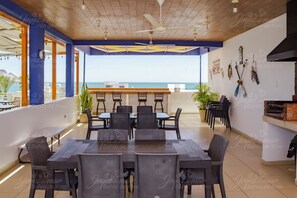 An outdoor dining area with a view of the sea, featuring a long table and chairs, a ceiling fan, and a brick oven.