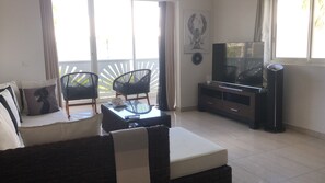 Un salon comprenant un canapé, deux fauteuils, une table basse en verre, un téléviseur posé sur un meuble en bois, et un balcon avec vue.