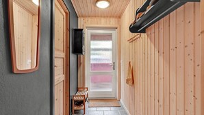 Ein Korridor mit Holzwänden, steinbelagtem Boden, einer Bank, einer Tür mit Frostglas und einem an der Wand montierten Fernseher.