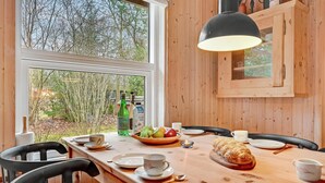 Ein Holztisch gedeckt mit Tellern, Tassen und einem Brot, mit Blick aus dem Fenster auf Bäume und Flaschen auf der Tischplatte.