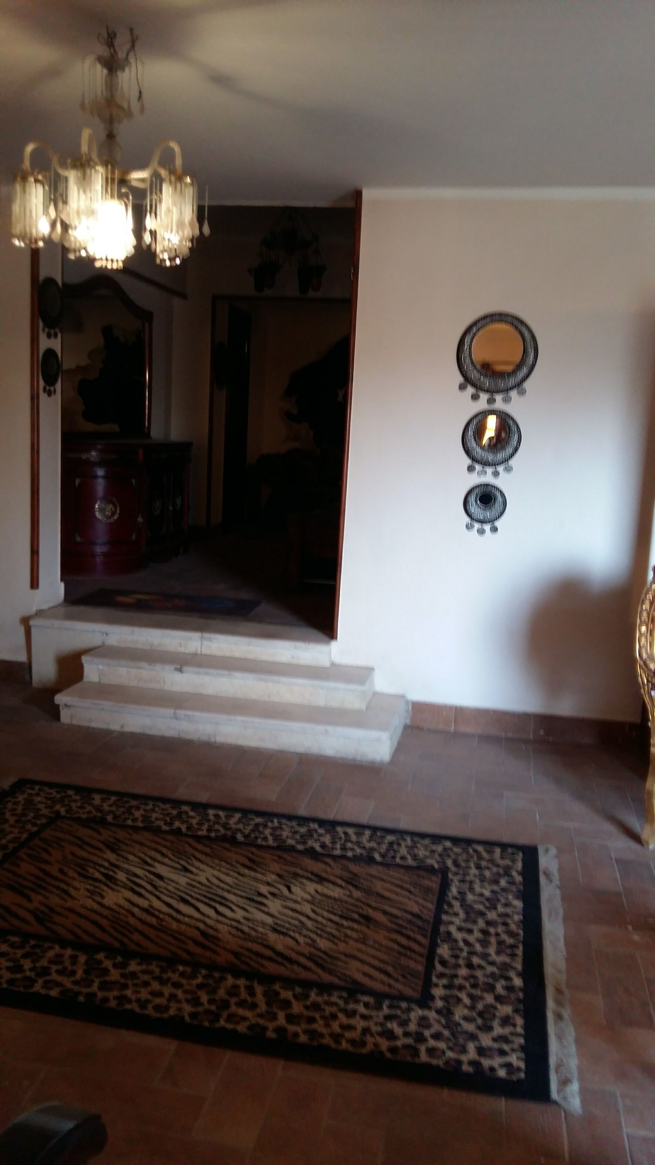 A room with a chandelier, a patterned rug, and a staircase leading to another area.