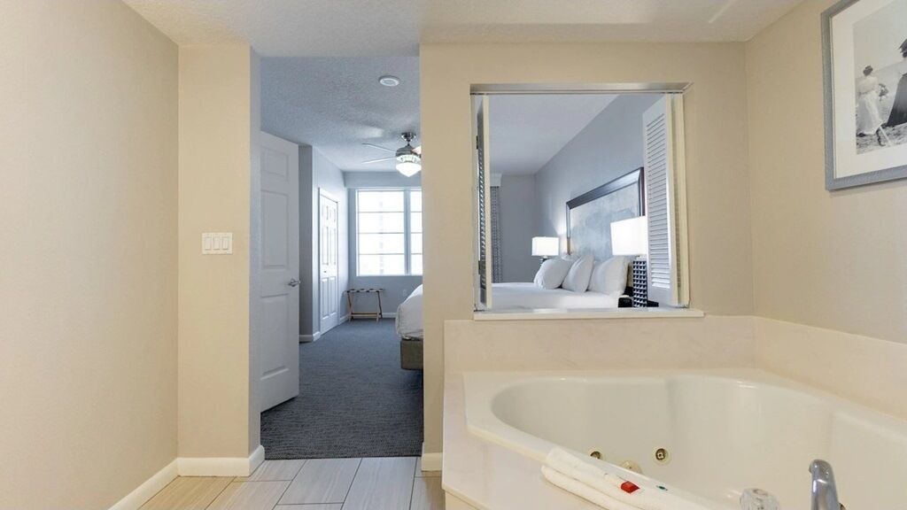 A modern bathroom with a large bathtub, a mirror, and a door leading to a bedroom.