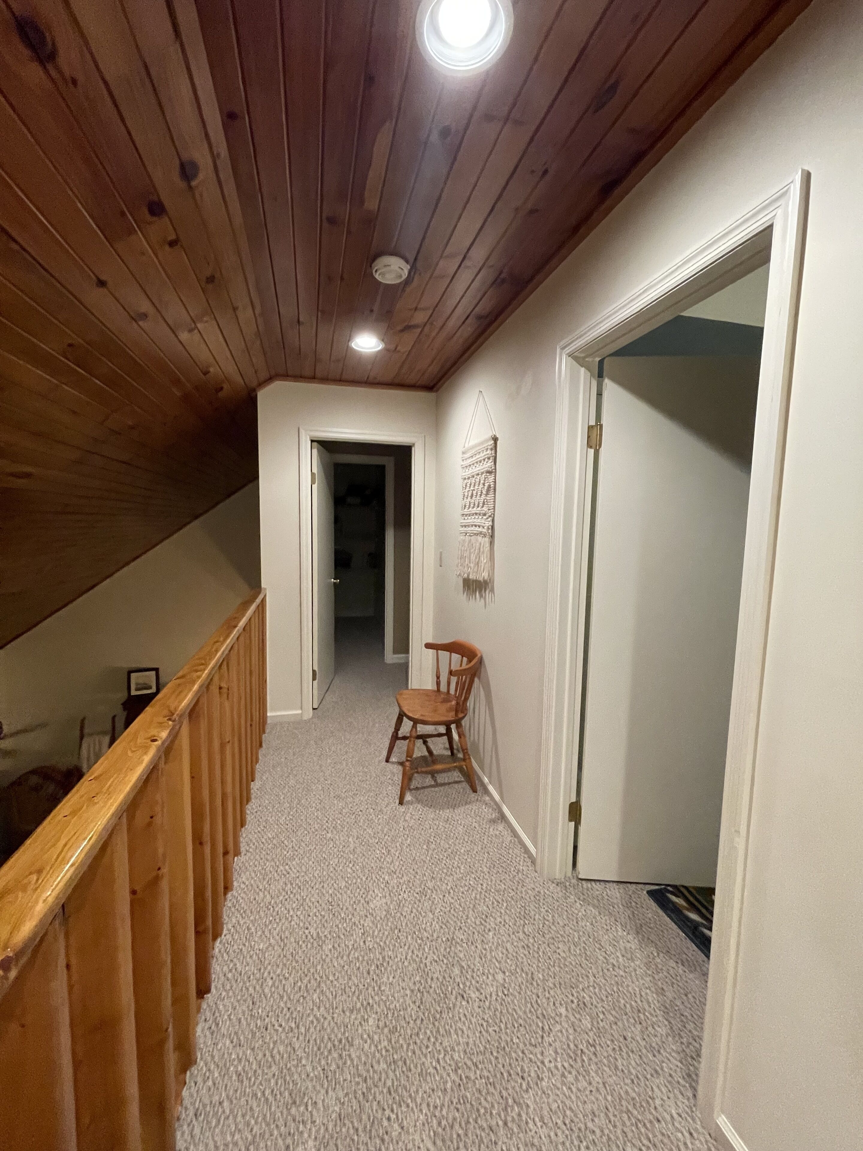 Een smalle gang met houten plafond en wanden, vloerbedekking, een houten stoel en een deur naar een andere kamer.