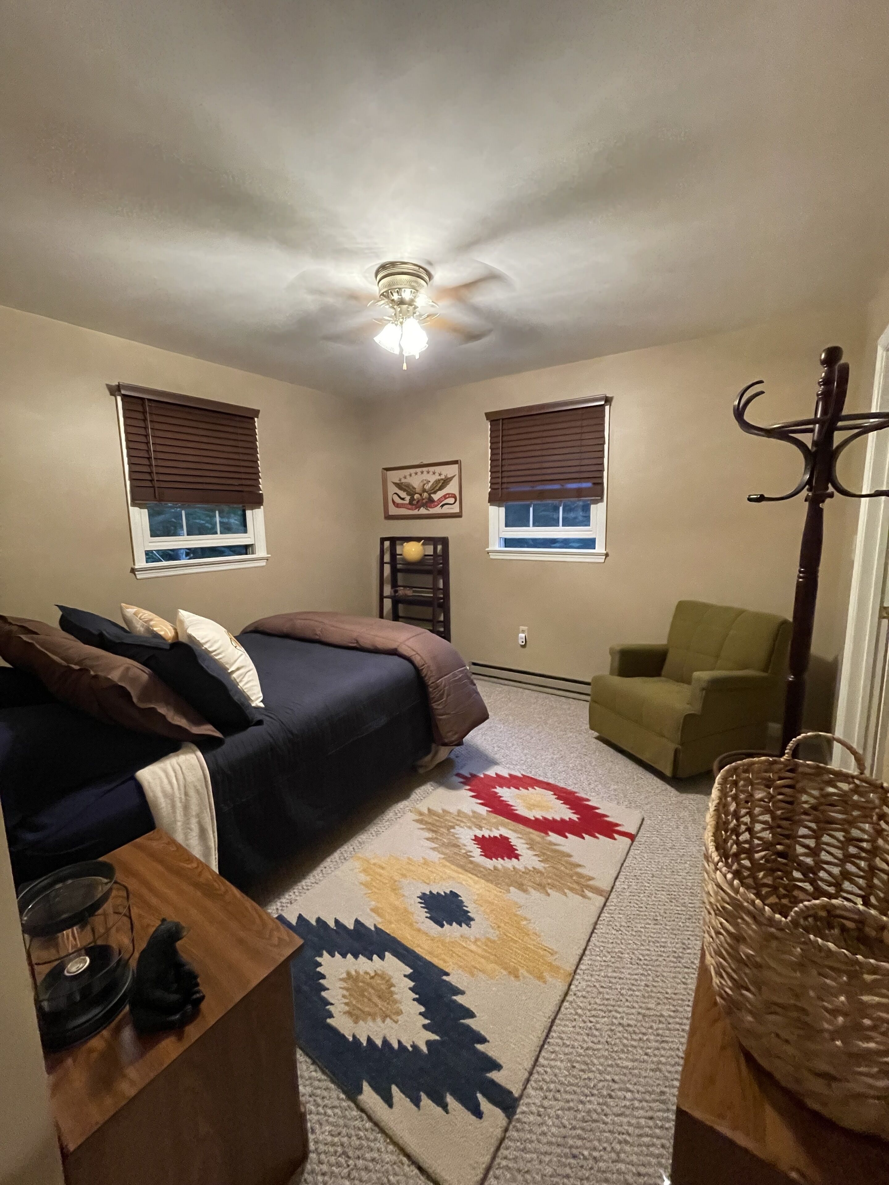 A bedroom with a bed, two windows with blinds, a ceiling fan, a green armchair, a woven basket, and a rug.