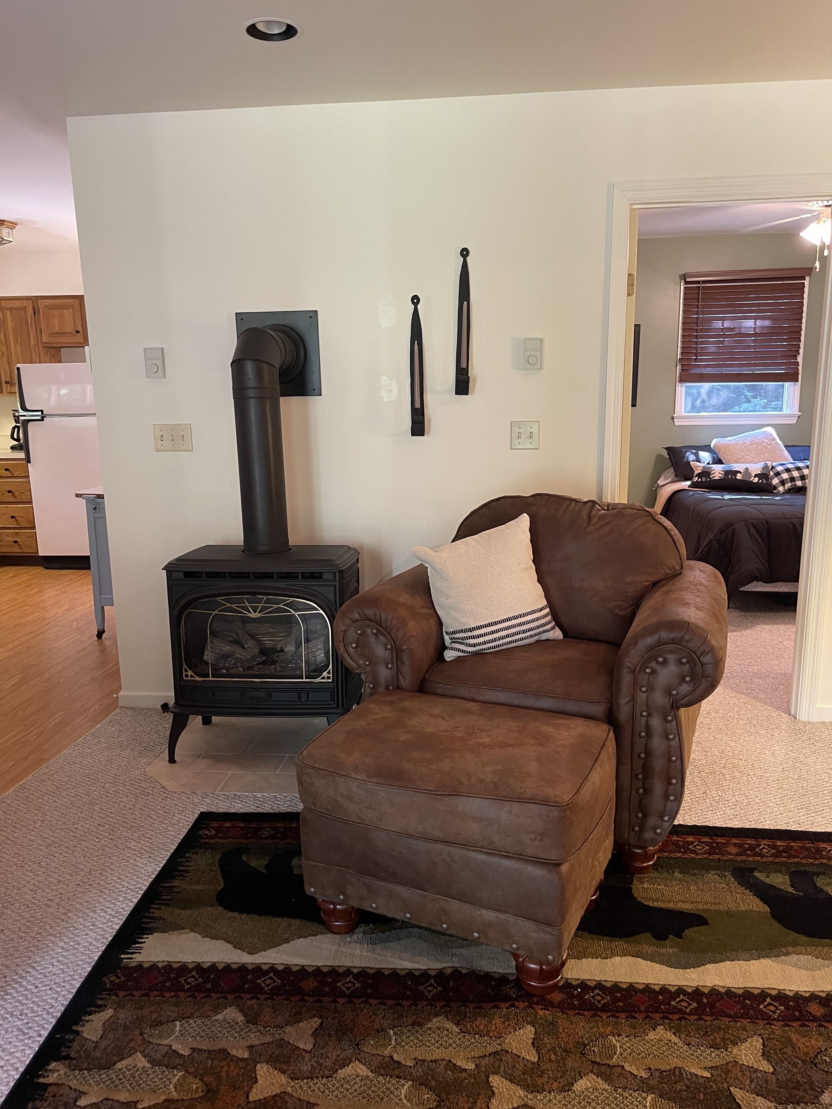 Een gezellige woonkamer met een leren fauteuil in bruin, een houtkachel in het zwart en een kamerkleed met een patroon.