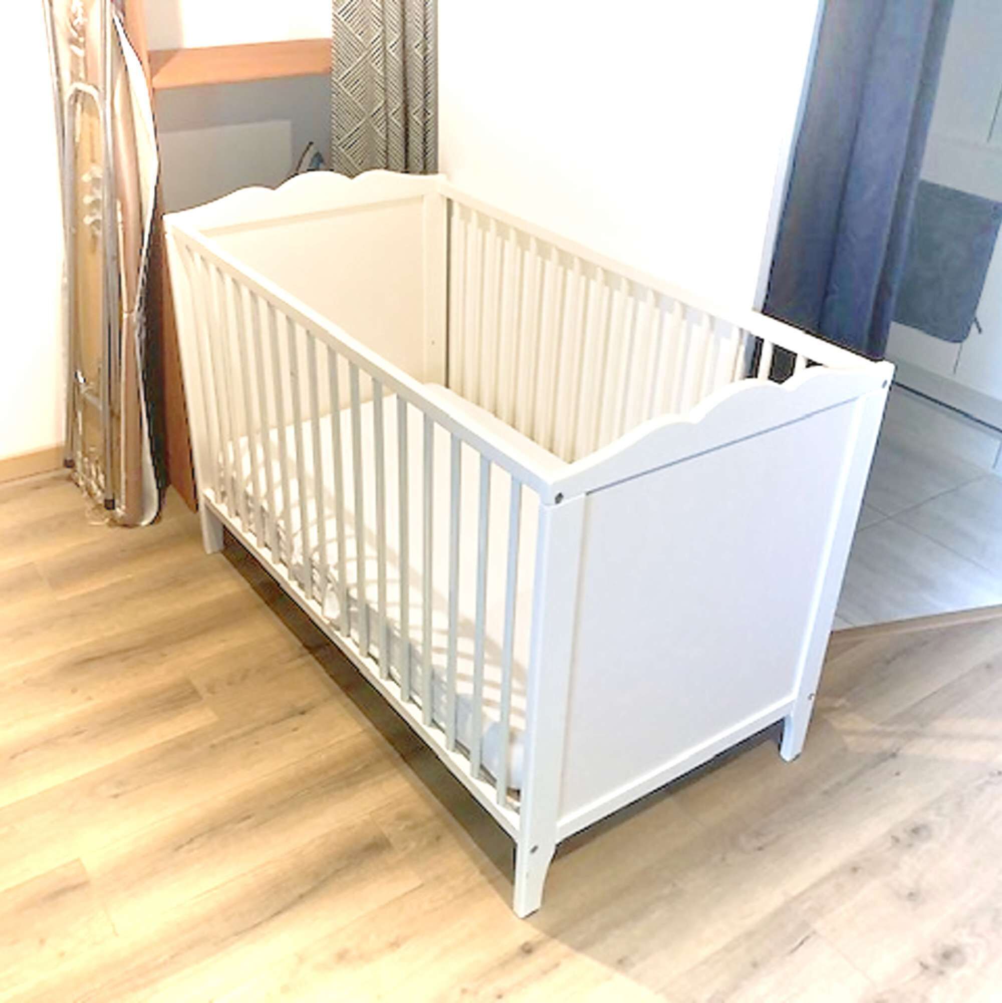 Un berceau blanc avec une barrière en métal, dans une pièce dont le sol est recouvert de planches de bois.