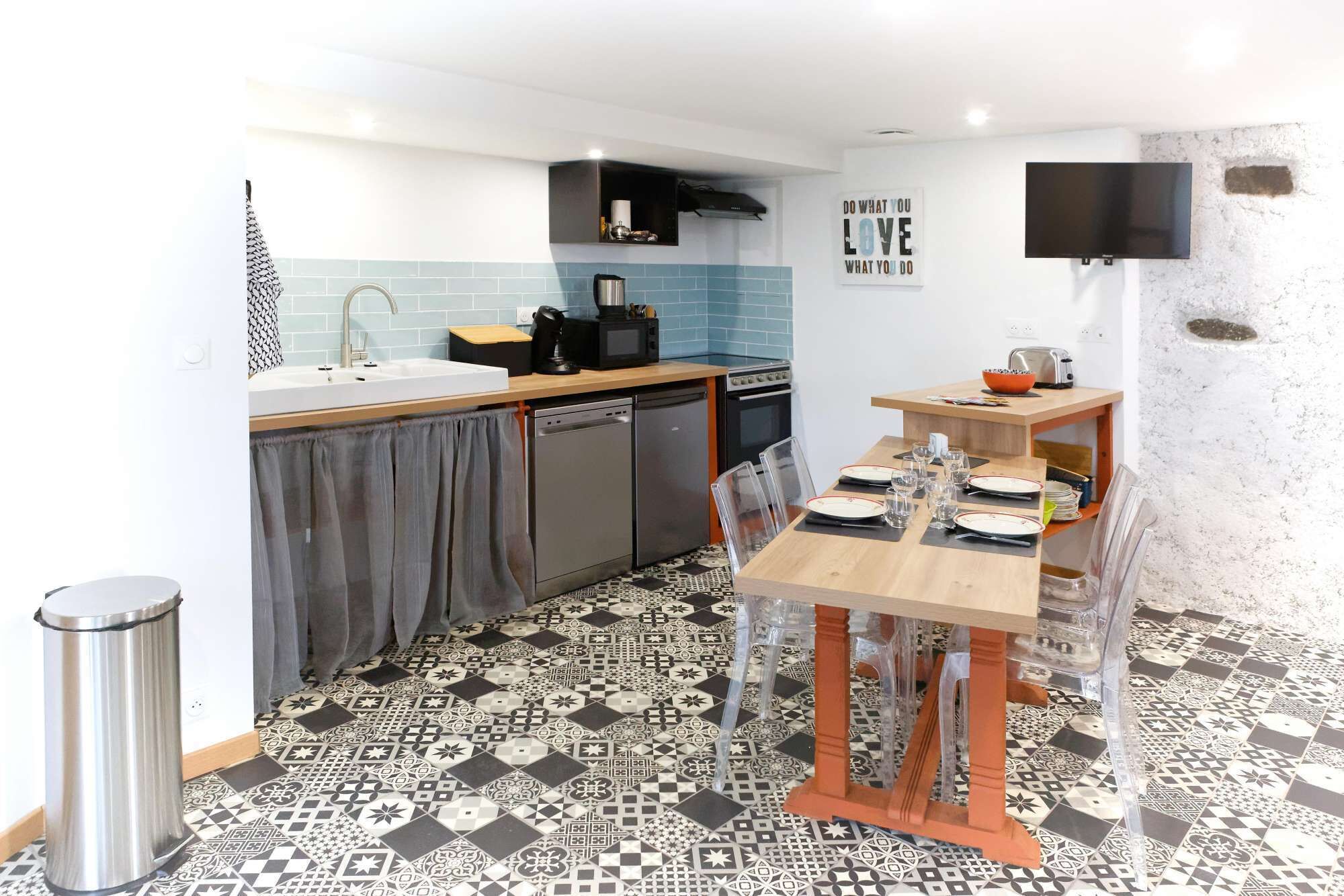 A compact kitchen with a sink, stove, and dishwasher, a dining table with chairs, and patterned floor tiles.