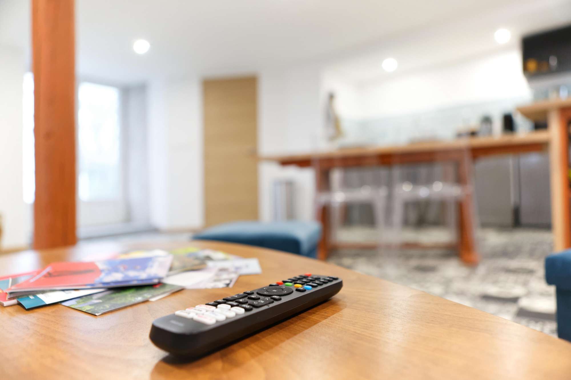 A remote control on a wooden table with magazines and a glass of water.