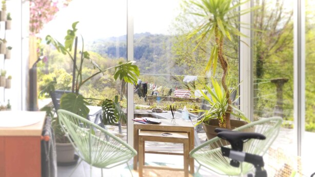 A glass-enclosed balcony with a wooden table, chairs, and potted plants.