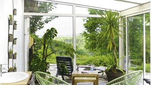 A conservatory with a glass ceiling, a wooden table, two green chairs, and potted plants.