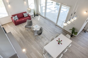 A modern living room with a red sofa, grey armchairs, a white dining table, and large windows.