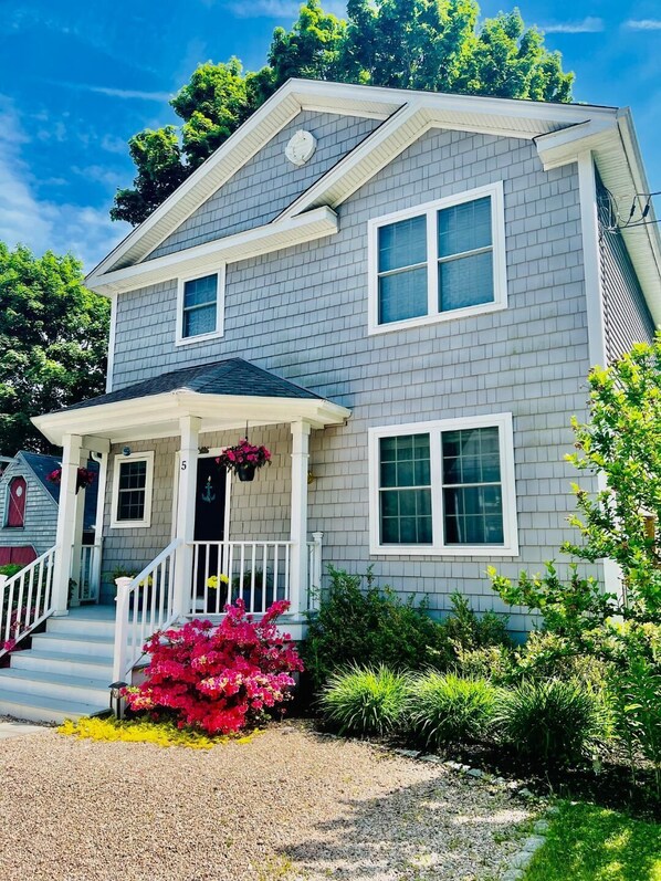 A two-story house with a front porch, a small garden with pink flowers, and a clear blue sky.