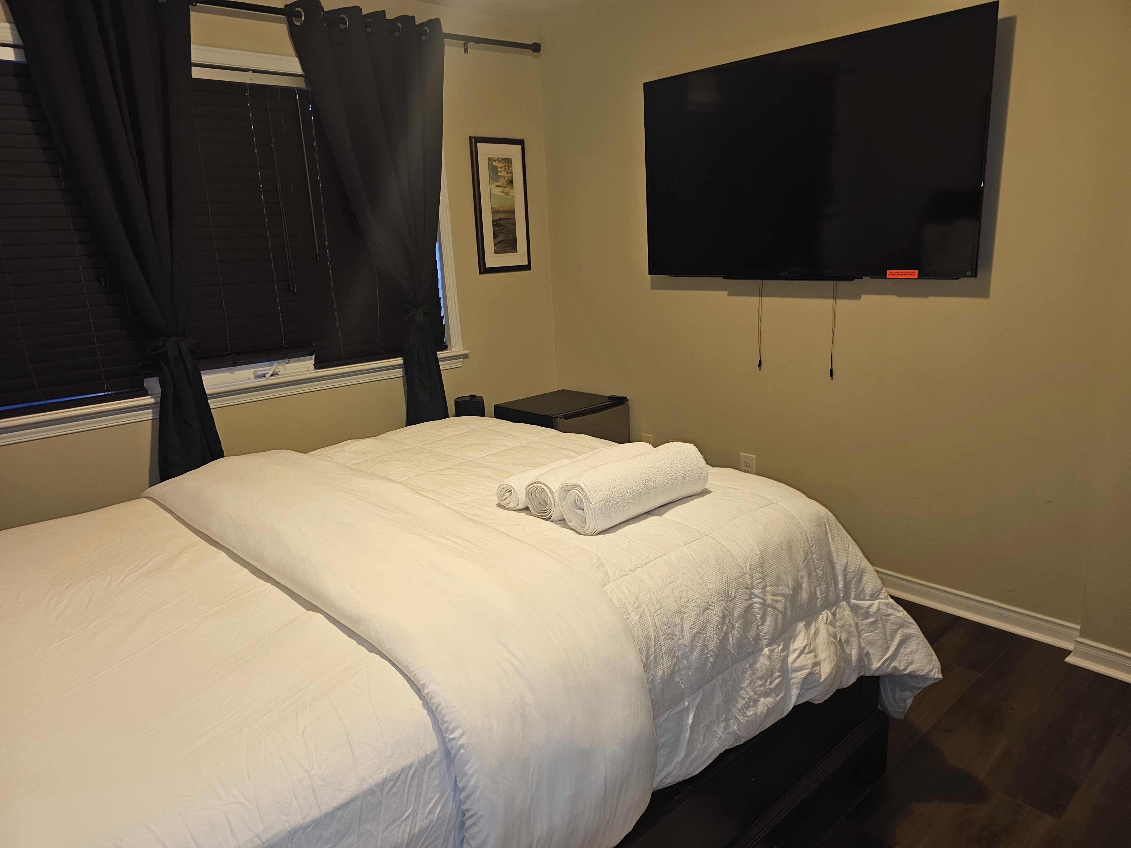 A neatly made bed with white bedding, a black TV mounted on the wall, and a window with black curtains.