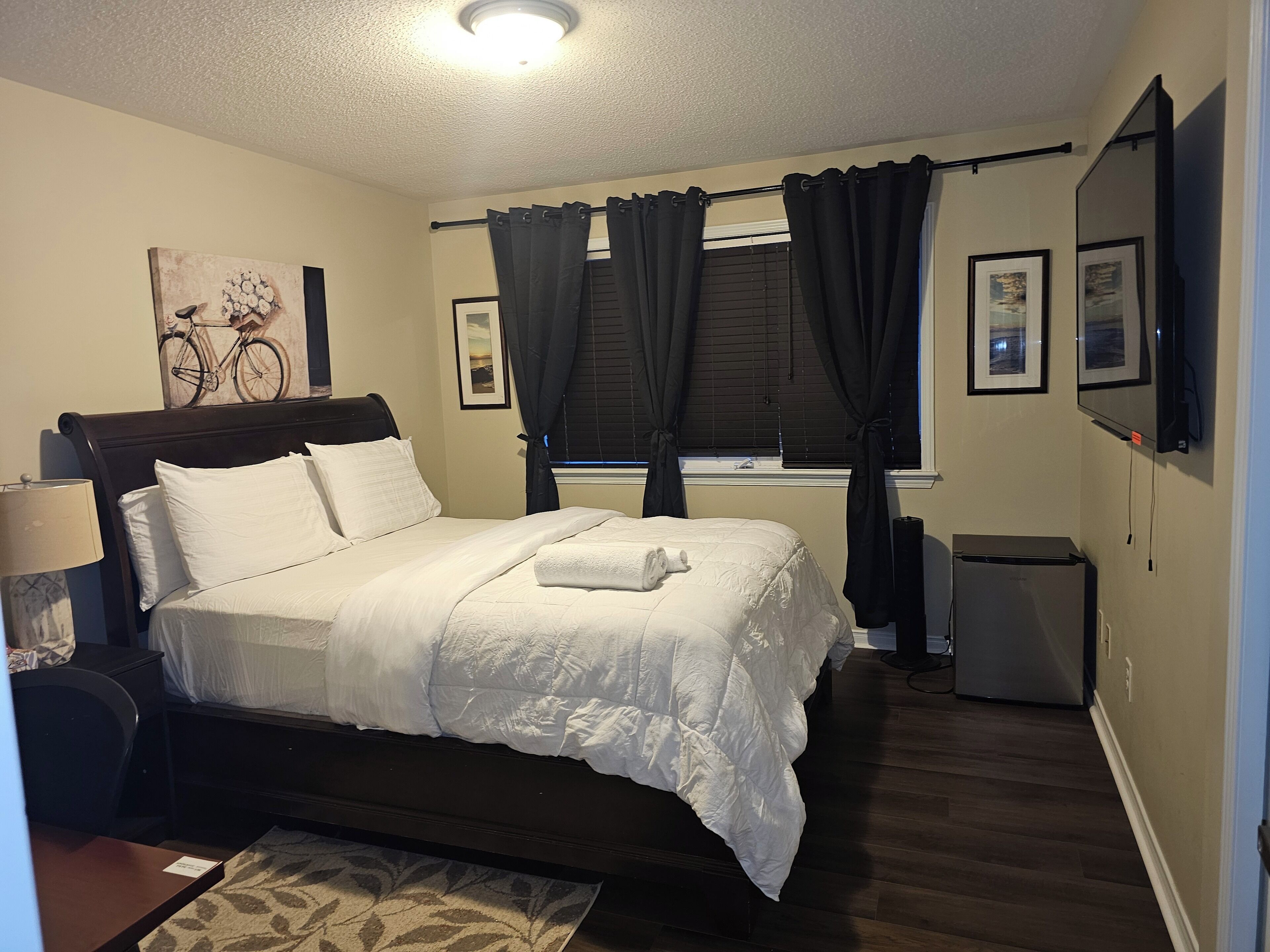 A neatly arranged bedroom with a bed, a bedside table, a lamp, a window with curtains, and a wall-mounted TV.