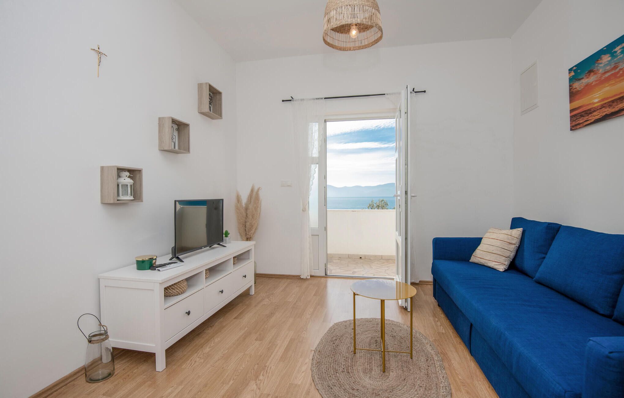Una sala de estar moderna con un sofá azul, un televisor sobre un mueble blanco y vistas al mar a través de una puerta abierta.