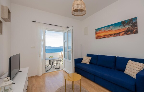 A living room with a blue sofa, a TV, a small table, and a painting of a sunset. - 1 bedroom beautiful apartment in Podaca (Podaca)