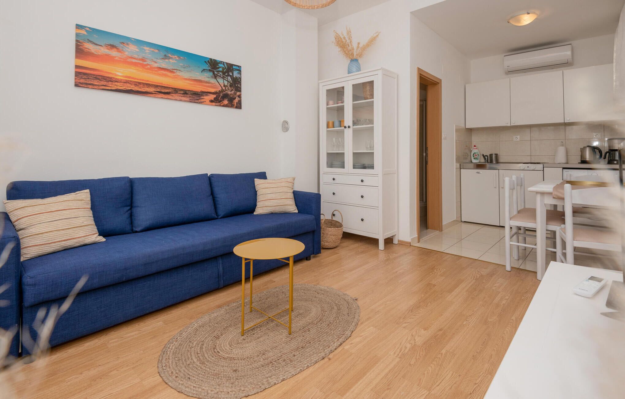 Een woonkamer met een blauwe bank, een klein rond tafeltje en een keuken met witte kasten en een koffieapparaat.